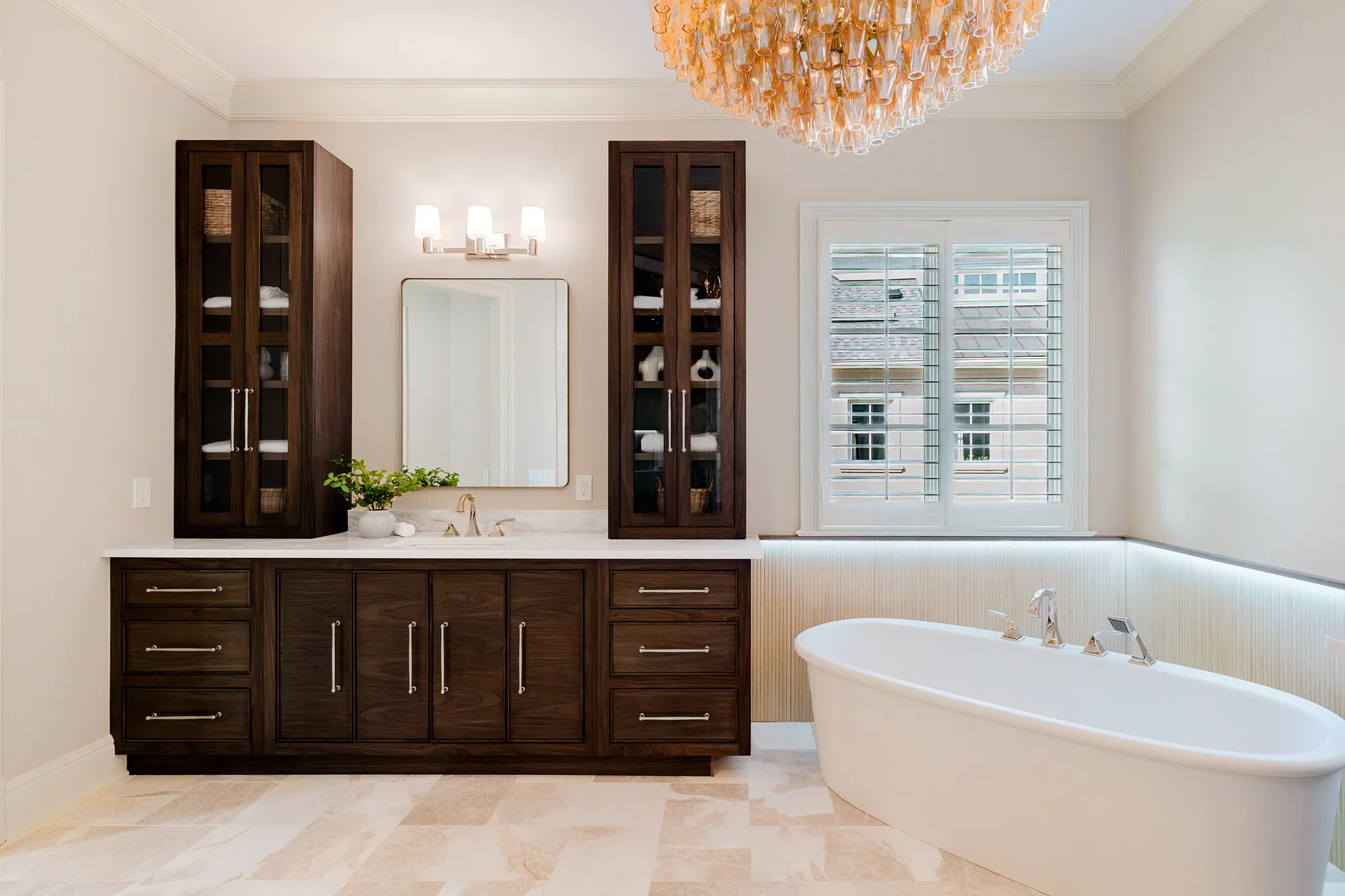 Primary bath with dark walnut cabinetry, a freestanding tub, and a crystal chandelier over travertine floors