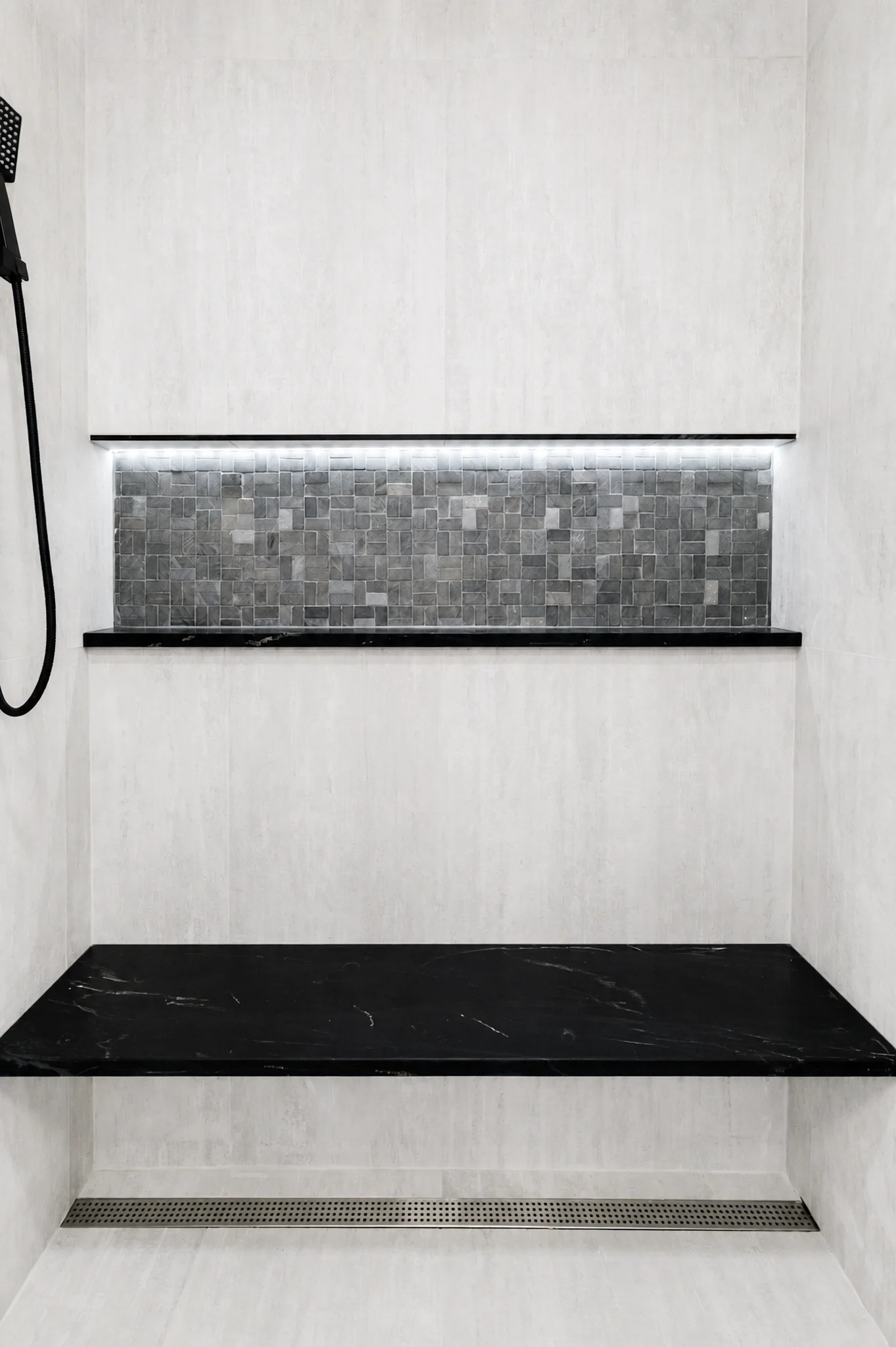 Illuminated shower niche with gray mosaic tile, black stone shelves, and a linear drain