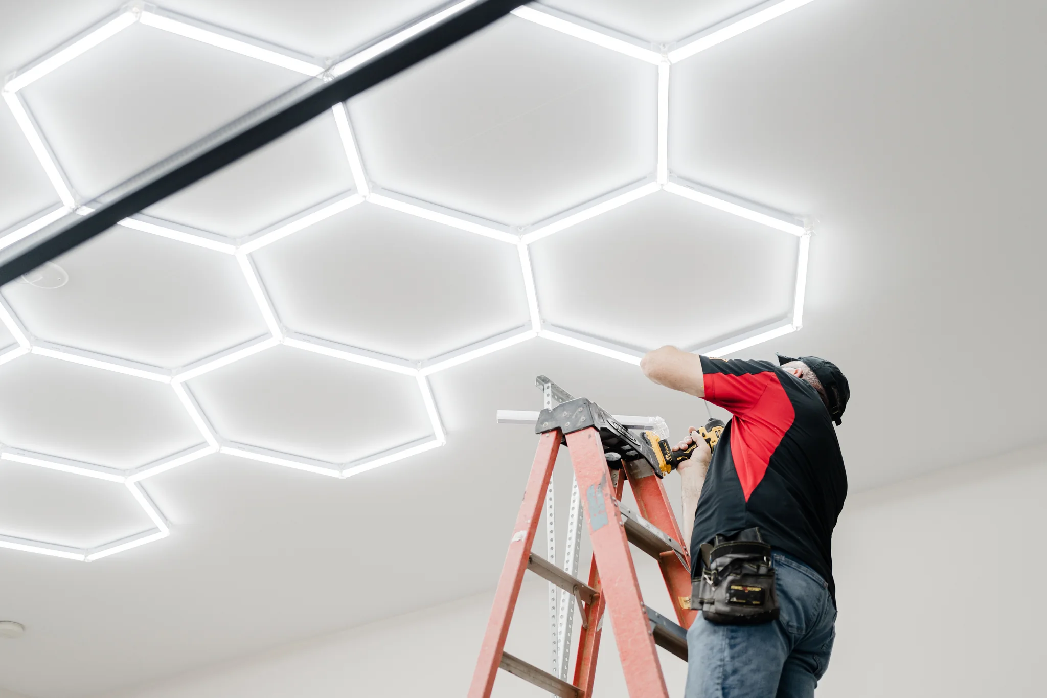 A Sharp crew member installing a custom geometric ceiling-light system from a ladder
